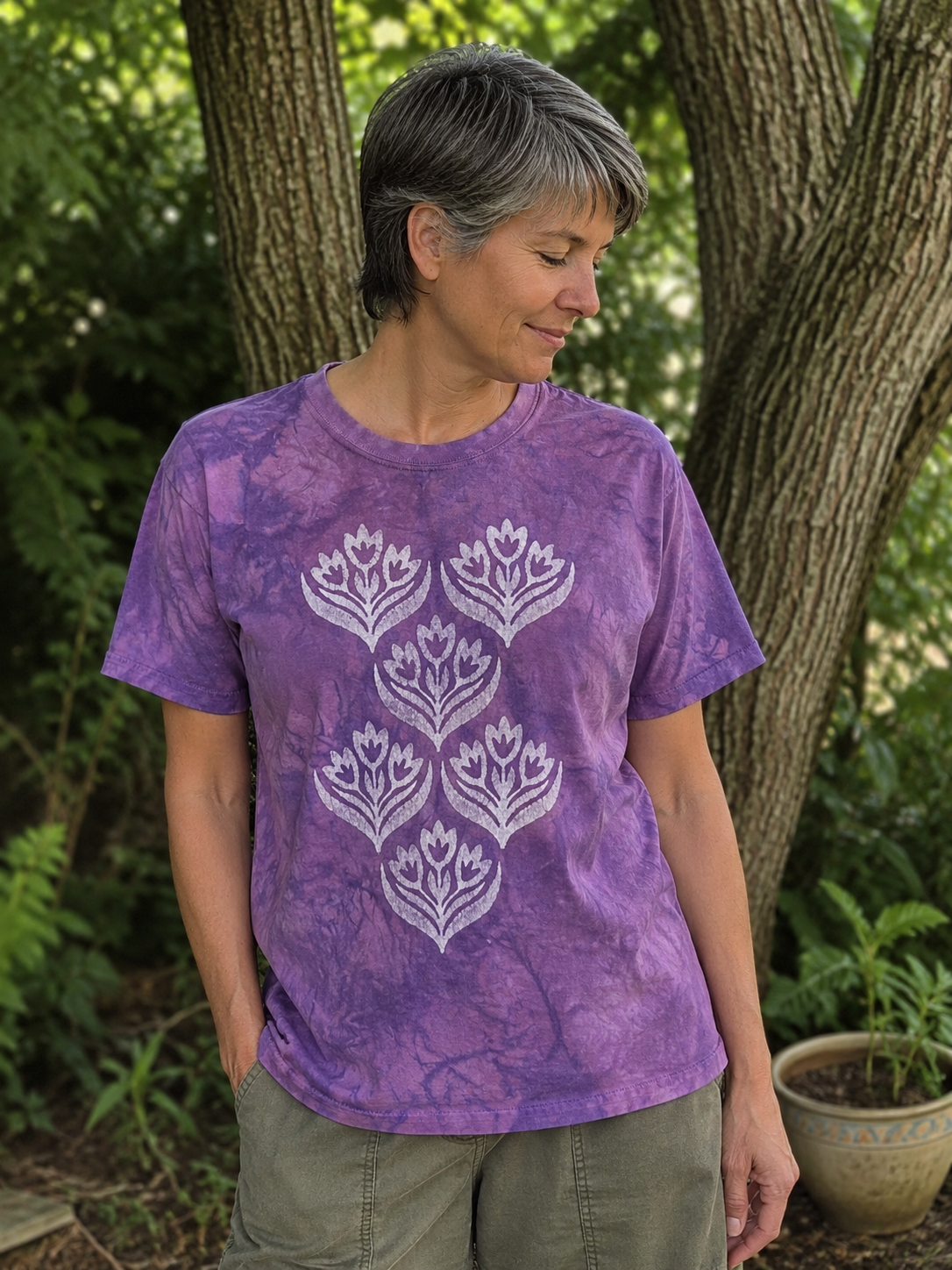 A woman wearing a purple short-sleeve t-shirt with a white floral block print design, standing outdoors in front of trees.